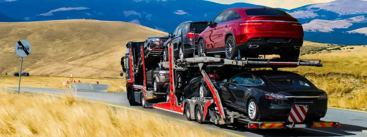 A red car carrier truck transports several cars against a scenic backdrop of birds flying in the distance, mountains, and a road sign.