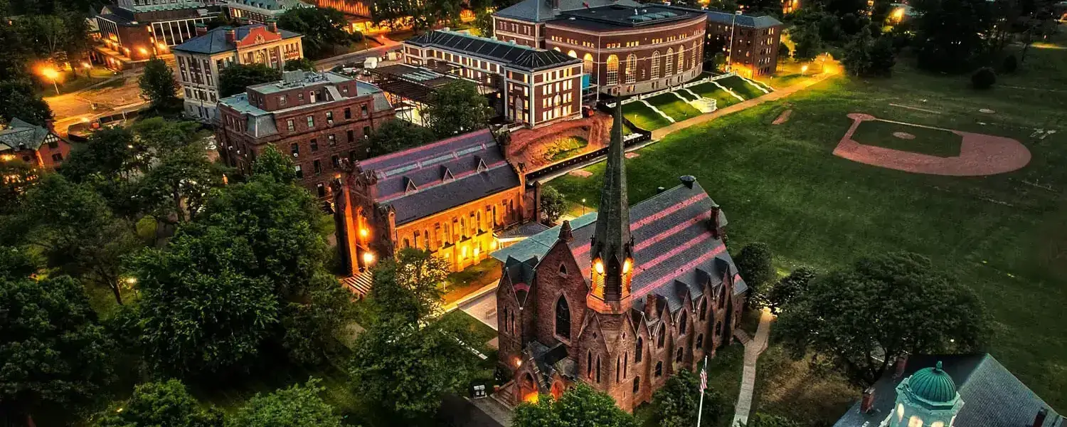 An aerial view of the Wesleyan University campus in Middletown, Connecticut, at night. The buildings are illuminated, with walkways and trees visible throughout the grounds