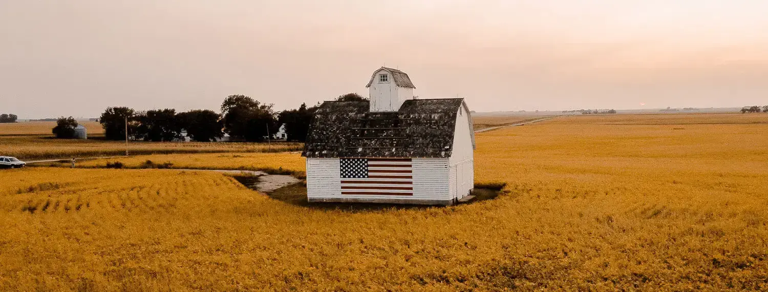 A weathered red barn with a large American flag painted on its side stands in a field of tall green cornstalks under a clear blue sky. This scene is typical of rural Iowa.