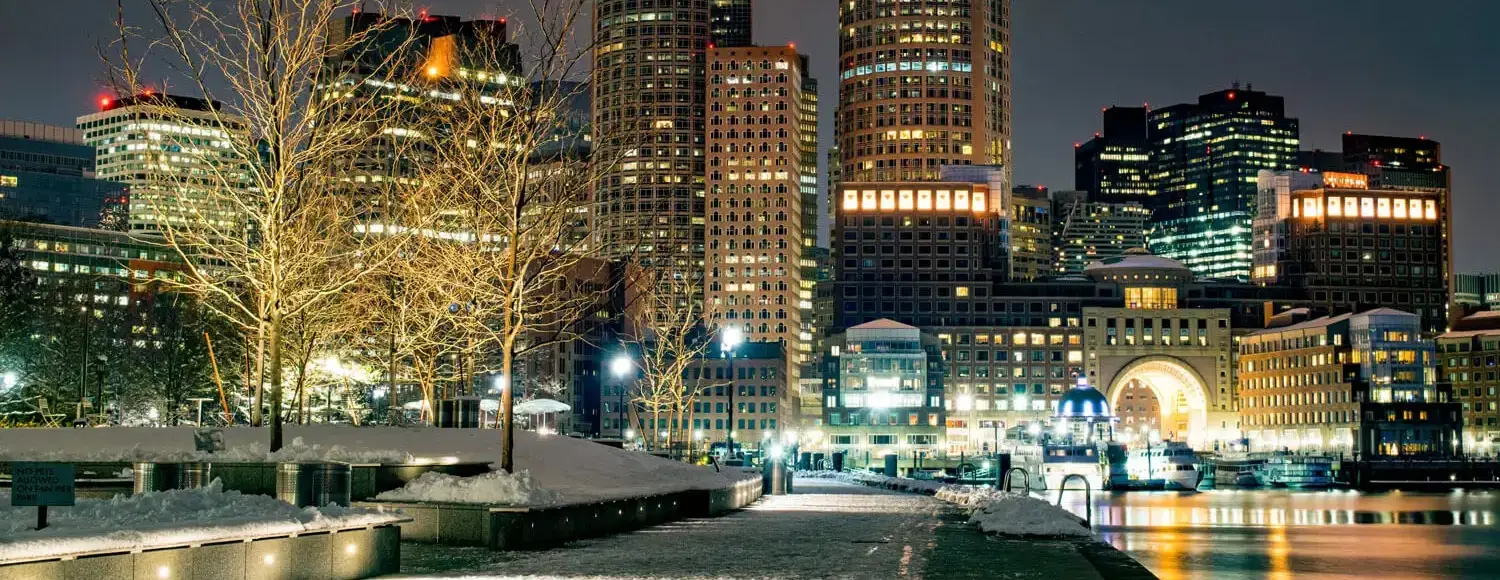 image of a city skyline at night. The buildings are illuminated with warm yellow lights, and there is a dusting of snow on the ground.