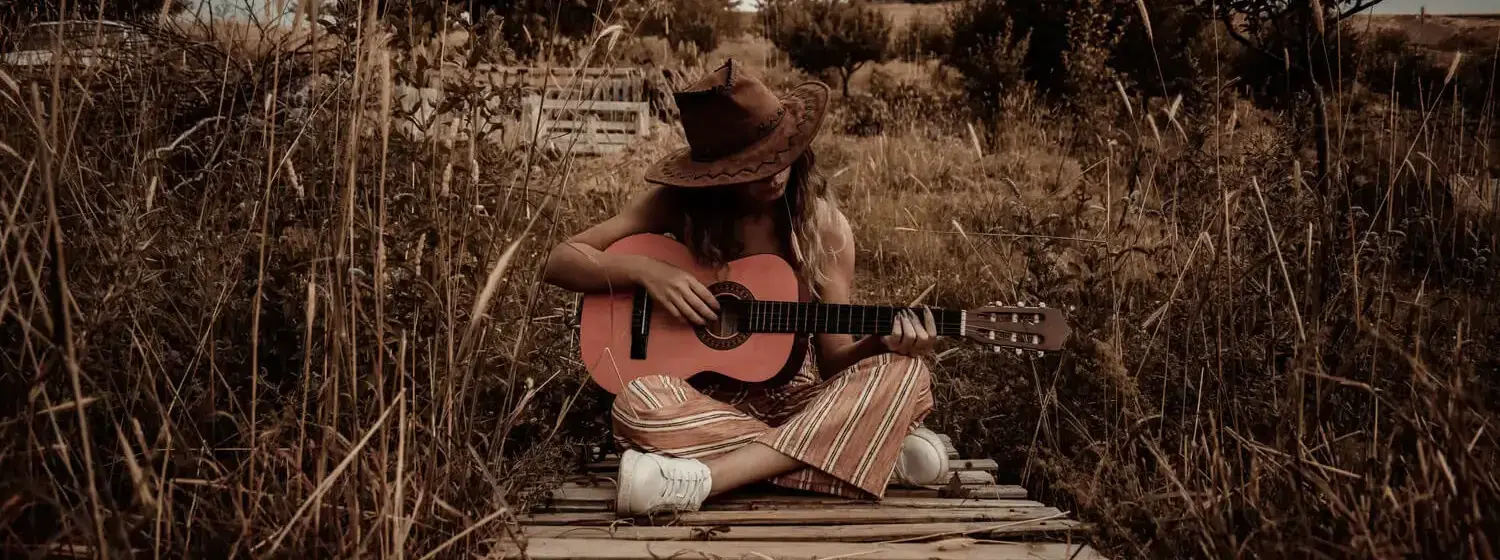 A person with a cowboy hat sitting in the middle of a field holding a guitar.
