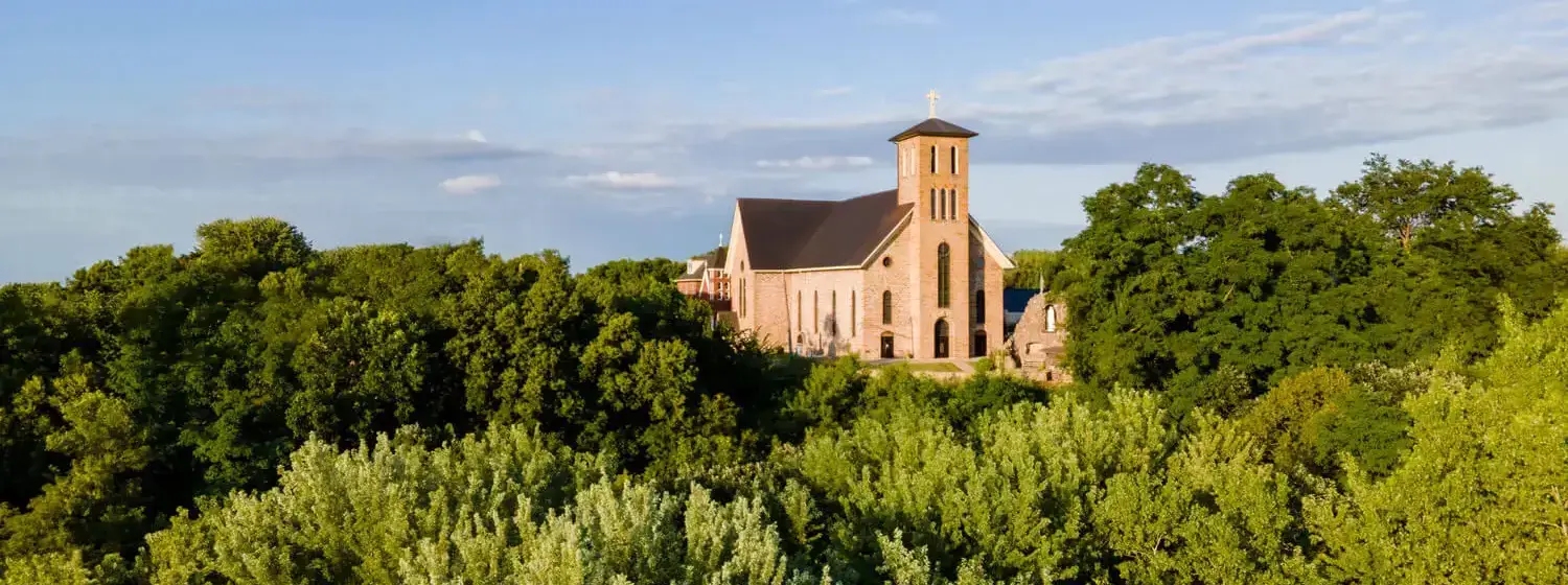 Aerial view of a church surrounded by trees. The church has a dark-colored roof and a cross atop of it.